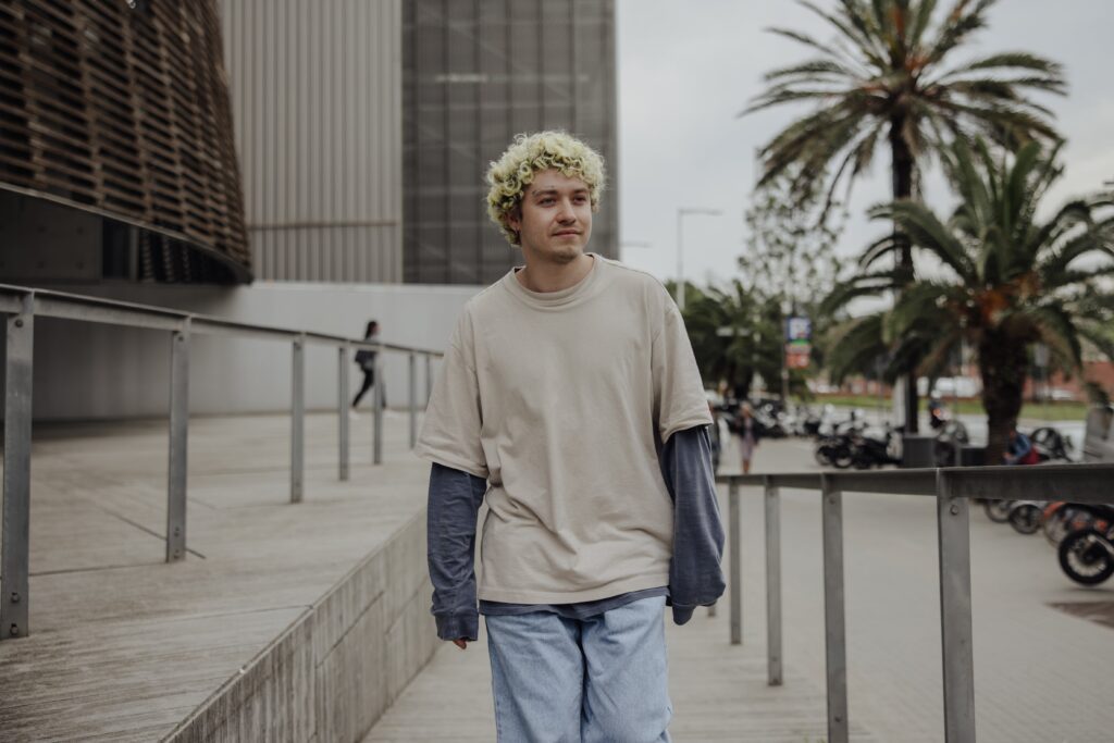 A imagem mostra um rapaz de cabelos loiros e encaracolados na rua. Ele veste uma camiseta manga longa azul por baixo de uma camiseta bege manga curta, ambas mais largas que o normal. Ele olha para o lado. No cenário da foto, à direita dele, vemos motos estacionadas e palmeiras.