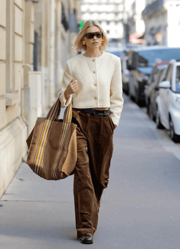 Na imagem há uma moça loira usando óculos escuro, um casaco de grife bege, uma bolsa grande marrom e uma calça marrom escuro. Ela está caminhando em uma calçada, com prédios em um lado e carros estacionados do outro.Na imagem há uma moça loira usando óculos escuro, um casaco de grife bege, uma bolsa grande marrom e uma calça marrom escuro. Ela está caminhando em uma calçada, com prédios em um lado e carros estacionados do outro.