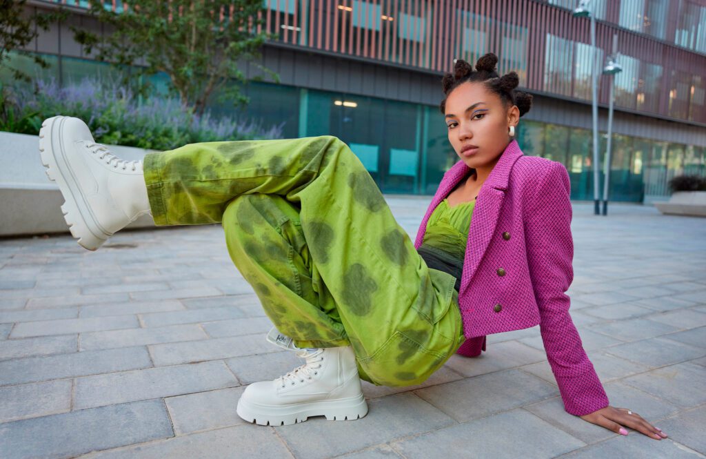Na imagem há uma jovem negra com cabelos presos em pequenos coques. Ela está em uma pose no chão, com as pernas cruzadas e as mãos tocando a calçada. Ela usa uma calça verde com pequenos manchas verde escuro, uma blusa verde e um blazer roxo por cima. Em seu pés há um coturno branco.